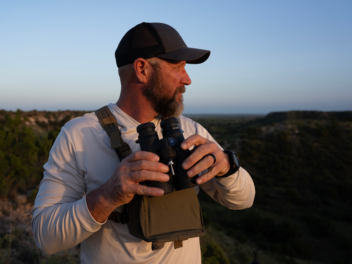 A hunter with a beard wearing a chest harness holds the lightweight ZEISS SFL 50 binoculars while scouting a valley during early morning light.