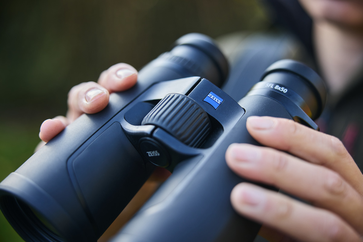 A close-up shot of a person's hands holding the ZEISS SFL 8x50 binoculars, emphasizing the SmartFocus wheel and the iconic blue ZEISS logo.