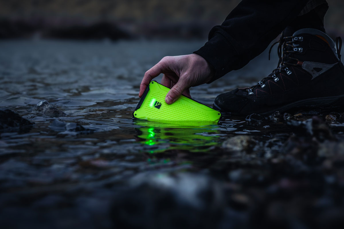 A hand placing a bright green, waterproof Dark Energy Poseidon Nano power bank into a rocky stream to demonstrate its IP68-rated durability.