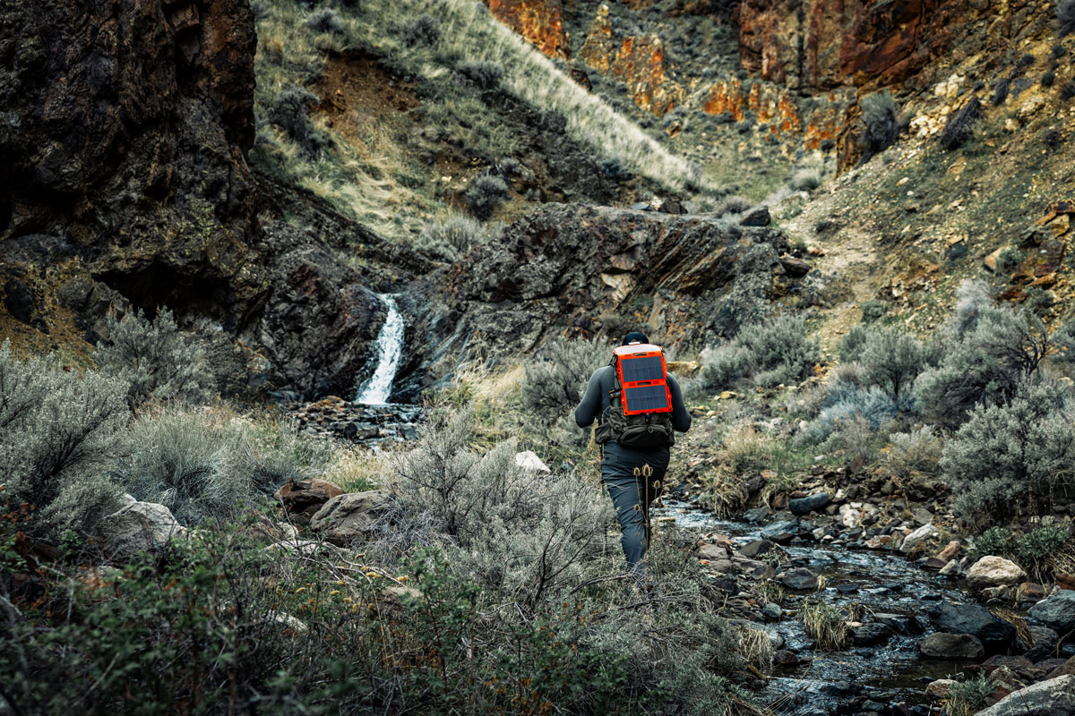A close-up of a hunter attaching a bright orange Dark Energy Spectre solar panel to his backpack in a forest setting.