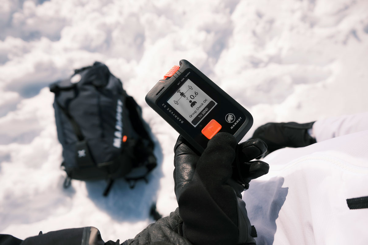 A gloved hand holds the Mammut Barryvox S2 avalanche transceiver, showing its clear MIP display with a 'Group Check Ok' message in a bright, snowy environment.