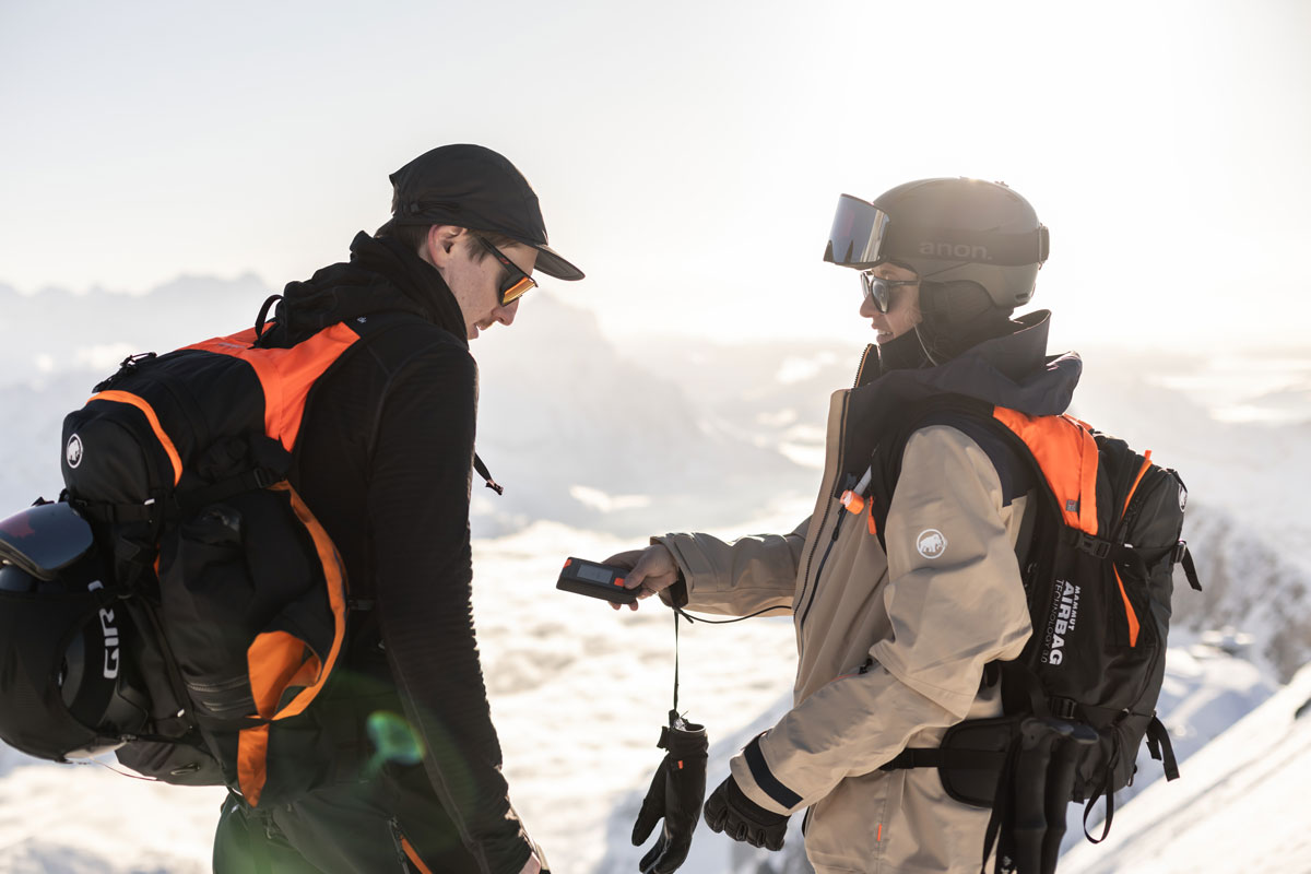 Two mountaineers in full ski touring gear use a Mammut Barryvox S2 transceiver on a sunny, snowy mountaintop, demonstrating avalanche safety training.